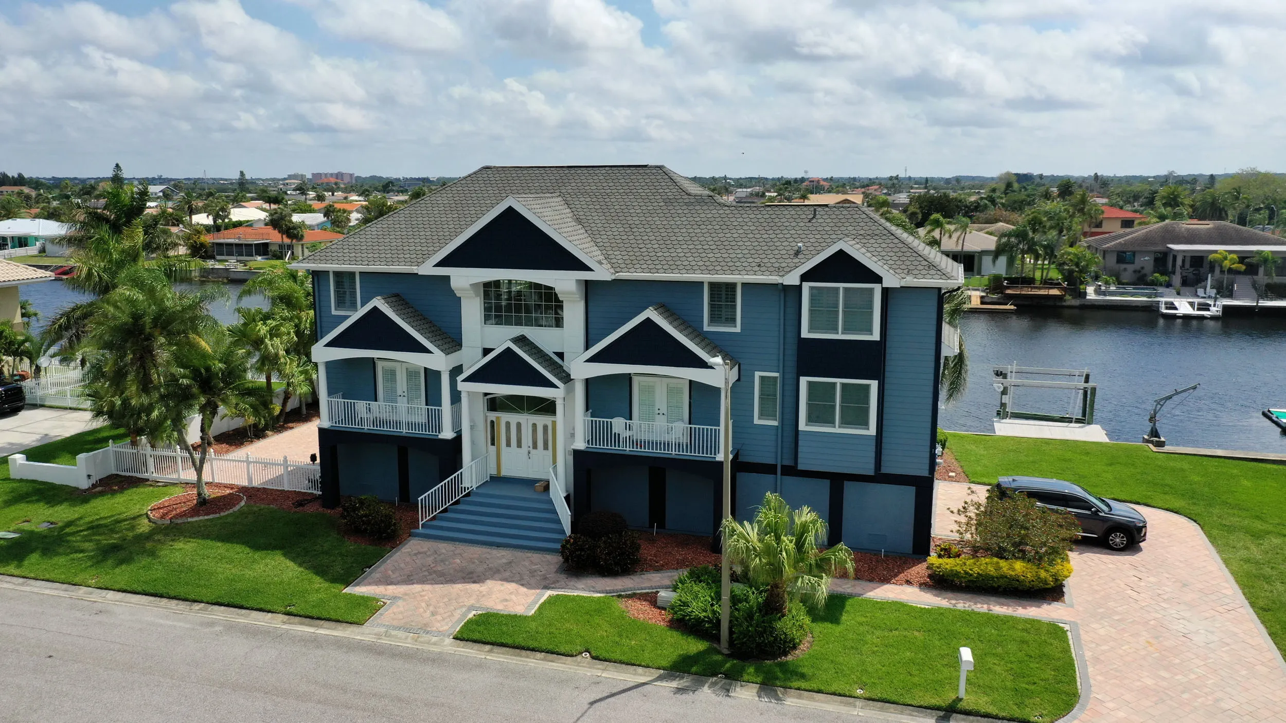 Blue two-story house with black and white trim, manicured lawn, driveway with a black car, and waterfront canal view.
